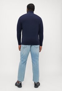 Homme debout de dos, portant un col roulé bleu marine, un jean bleu clair et des baskets noires, sur un fond clair uni.