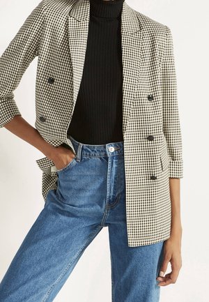 Personne portant un col roulé côtelé noir, un jean bleu taille haute et un blazer croisé à carreaux beige et noir aux manches retroussées.