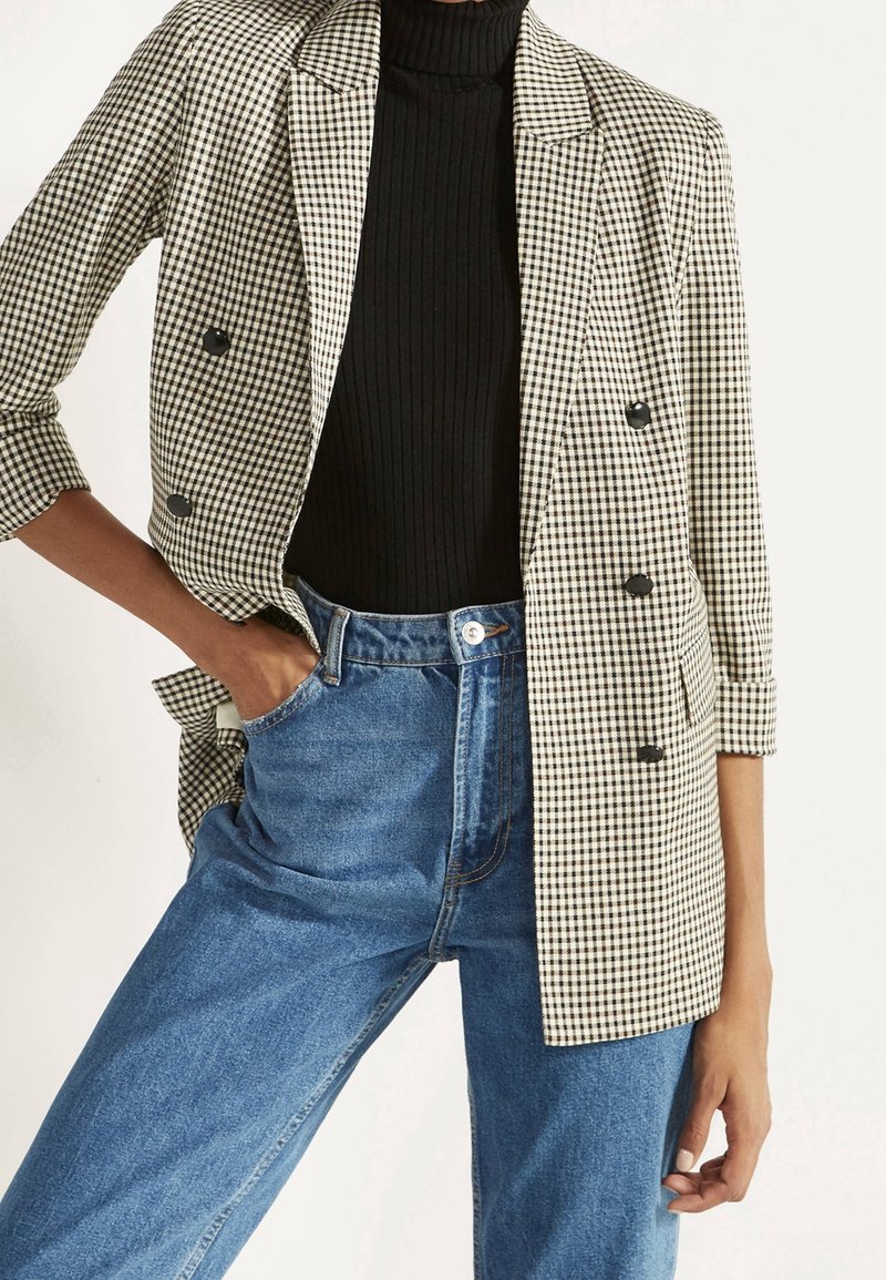 Personne portant un col roulé côtelé noir, un jean bleu taille haute et un blazer croisé à carreaux beige et noir aux manches retroussées.