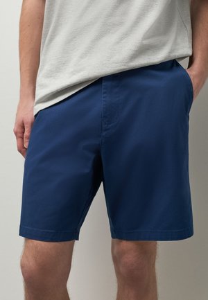 Mężczyzna ubrany w jednolite granatowe szorty i jasnoszary t-shirt, z jedną ręką w kieszeni, a drugą spoczywającą wzdłuż ciała.