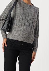 Woman wearing a gray ribbed sweater, black pants, and carrying a black shoulder bag against a white background.