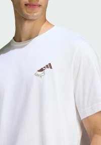 Biela bavlnená tričko s malou hnedou grafikou kávového hrnčeka s tekutinou striekajúcou von, krátke rukávy a okrúhly výstrih.