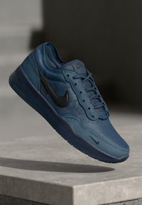 Tumedast sinised Nike spordijalatsid tekstureeritud pealispinnaga, musta logoga, ümmarguse varbaga, paeltega ja pehme tallaga, hõljuvad neutraalse taustaga.