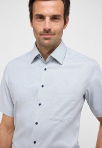 Camicia azzurra a maniche corte con bottoni, con un sottile motivo a griglia, dotata di una tasca sul petto e bottoni blu scuro.