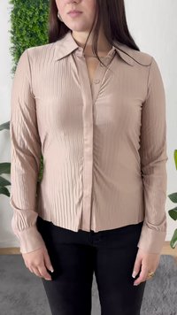 Frau in einer hellrosa, gefältelten Bluse mit langen Ärmeln und schwarzen Hosen, die drinnen neben grünen Pflanzen auf einem grauen Teppich steht.
