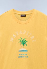 Kollane puuvillane T-särk ümmarguse kaelusega. Omab graafilist trükist palmipuude ja tekstiga "NAPAPIJRI RIVIERA LIFESTYLE" valges ja sinises värvitoonis.