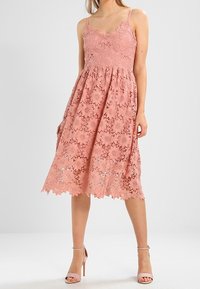 Robe en dentelle rose pâle avec des bretelles spaghetti, un motif floral et un ourlet festonné, dotée d'un corsage ajusté et d'une jupe évasée.
