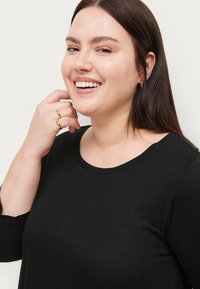 Sort langærmet top med en rund halsudskæring. Glat stof med en let glans. Modellen har hånden på hagen og bærer en guldring.