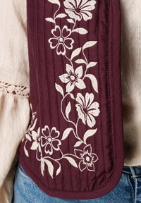 Bordeauxrode vest met witte bloemrijke borduursels, gestructureerde stof, met verticale lijnen en een afgeronde zoom, gedragen over een lichtgekleurde top.