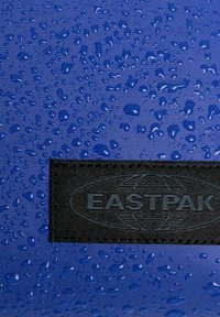 Blått tyg med vattenpärlor. Svart texturerad etikett med "EASTPAK"-logotypen längst ner. Materialet verkar vara vattenavvisande.