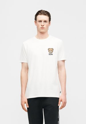 Ung mand med bagstrøget hår iført en hvid T-shirt med en lille teddybjørn-logo og teksten "Moschino Underwear" på brystet.