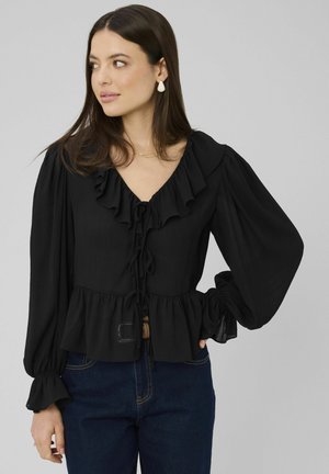 Femme portant un chemisier noir à volants avec des manches longues bouffantes et un jean bleu foncé, regardant sur le côté sur un fond uni.