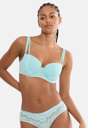 Kobieta z plecionymi włosami, ubrana w jasnoniebieski biustonosz wykończony koronką i dopasowane majtki, stojąca z jedną ręką na biodrze na jednolitym tle.