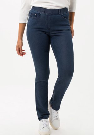 Kvinde iført mørkeblå slim-fit jeans, hvide sneakers og en hvid langærmet top, stående mod en ensfarvet baggrund.