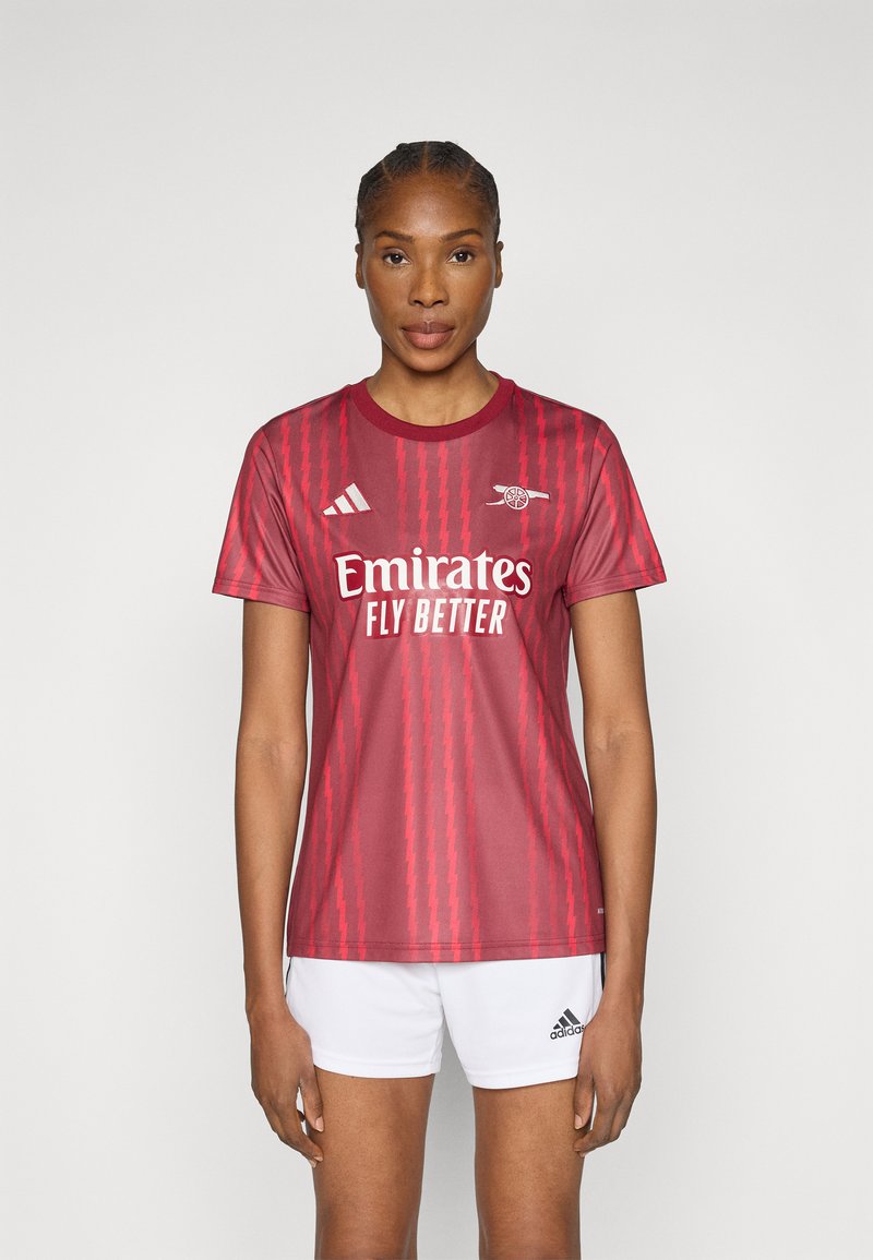 Κόκκινη αθλητική φανέλα με κοντά μανίκια και στρογγυλή λαιμόκοψη, με διαγώνιες ρίγες και λευκό λογότυπο. Φορεμένη με λευκά σορτς.