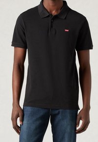 Czarna bawełniana koszulka polo z klasycznym kołnierzykiem, trzema guzikami oraz małym czerwonym logo Levi's po lewej stronie klatki piersiowej. Noszona z niebieskimi dżinsami.