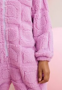 Une combinaison rose floue avec un motif en blocs texturé, dotée d'une fermeture éclair à l'avant et de manches resserrées. Tissu doux et pelucheux avec une apparence volumineuse.