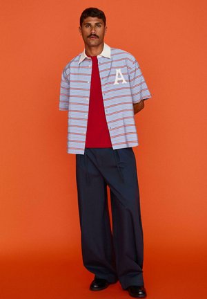 Bajszos férfi kék-piros csíkos rövid ujjú inget visel piros póló felett, sötét, bő szárú nadrágot és fekete cipőt, narancssárga háttér előtt áll.