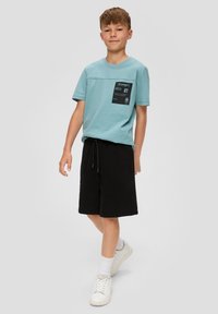 Lichtblauwe katoenen t-shirt met korte mouwen en een voorzakje, gecombineerd met zwarte shorts met koord en witte sneakers.