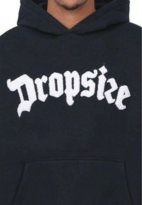 Crna dukserica s prednjim dzepom, s bijelim, pahuljastim izvezenim tekstom "Dropskie" u podebljanom, gotičkom fontu. Mekana tekstura tkanine.