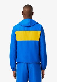 Blauwe hoodie met een grote gele streep over de achterkant. Gemaakt van glad materiaal met elastische manchet en zoom.