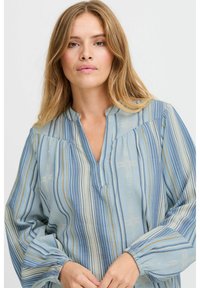 Blusa azzurra chiara con righe verticali bianche e dorate. Caratterizzata da un collo a V, dettagli arricciati sulle spalle e maniche ample.