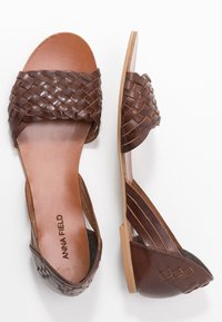 Sandales plates en cuir marron avec des lanières tressées sur les orteils et le talon arrière, étiquetées "ANNA FIELD", vues de dessus et de côté.