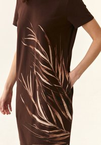 Brown short-sleeve dress featuring a large, abstract cream and dark brown leaf print on the front, with a side pocket. Soft cotton material.