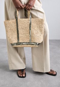 Wykonana z plecionej słomy torba na ramię w zielonych metalicznych akcentach i uchwytach, o prostokątnym kształcie i teksturowanej powierzchni, trzymana przez osobę.