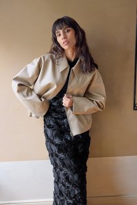 Chaqueta cropped beige con un patrón texturizado y gran collar, llevada sobre un vestido negro con aplicaciones de rosa en 3D.