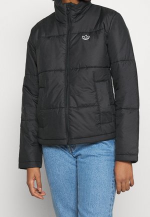 Personne portant une veste matelassée noire à fermeture éclair avec un petit logo Adidas blanc et un jean en denim bleu, debout devant un fond uni.