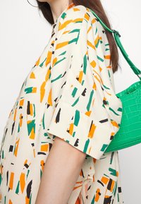 Camisa crema con patrones abstractos multicolores en verde, naranja y negro. Un bolso verde brillante está colgado del hombro.