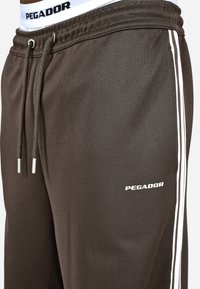 Braune Jogginghose aus weichem Material mit weißen Seitenstreifen. Verfügt über einen elastischen Bund mit Kordelzug und ein kleines "PEGADOR"-Logo.