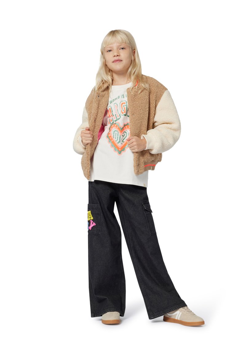 Blond meisje in een tan en crème fleecjas over een wit T-shirt met opdruk, zwarte wijde broek met patches en beige sneakers.