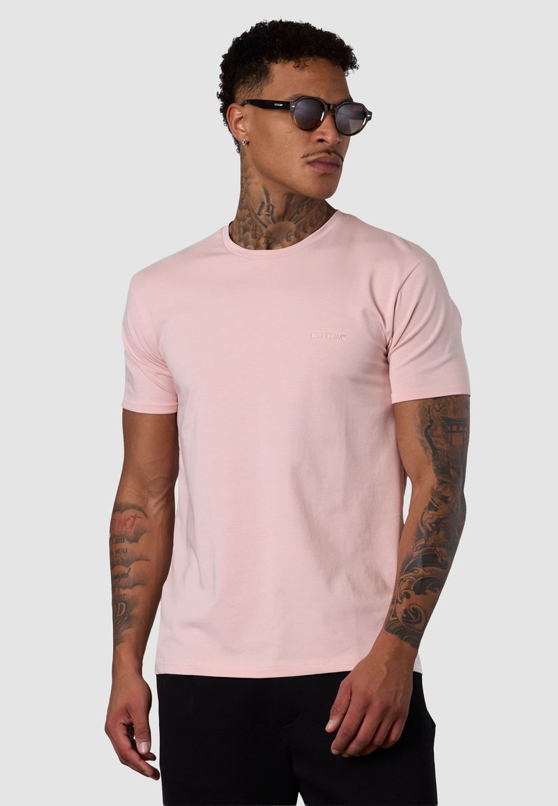 T-shirt rose clair à manches courtes avec col rond et détail de logo discret sur la poitrine. Tissu lisse, coupe standard, porté avec des lunettes de soleil noires.
