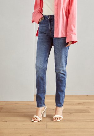 Camicia oversize rosa chiaro con bottoni sopra un top bianco, abbinata a jeans blu a vita alta e sandali bianchi con tacco a blocco.