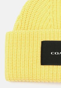 Gorro de punto amarillo con textura acanalada y dobladillo. Presenta una etiqueta rectangular negra con texto blanco.