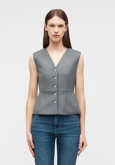 Γυναίκα που φορά αμάνικο γκρι τοπ με V λαιμόκοψη και πέντε διακοσμητικά κουμπιά, συνδυασμένο με μπλε τζιν, στέκεται μπροστά σε απλό φόντο.