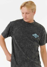 Camiseta de algodón negra, lavado, con mangas cortas. Cuenta con un pequeño logo azul en el lado izquierdo del pecho. Apariencia texturizada y ligeramente desvaída.