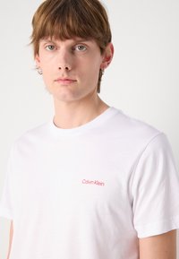 Hvit bomulls t-skjorte med rund hals og korte ermer. Har en liten rød brodert "Calvin Klein"-logo på venstre brystområde.