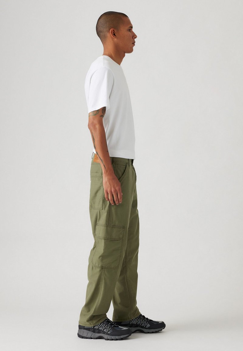 Pantaloni cargo verdi con molte tasche, realizzati in tessuto misto cotone. Abbinati a una t-shirt bianca a maniche corte e a sneaker nere con texture.
