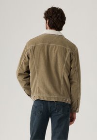 Veste en velours côtelé beige avec un col en fourrure blanche, présentant une coupe droite, des poignets boutonnés et des poches latérales. La vue arrière met en valeur le tissu texturé.