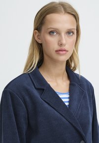 Veston en velours côtelé bleu marine avec un col à revers et un tissu texturé. Comprend un haut rayé bleu et blanc en dessous.