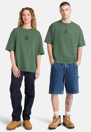 Due modelle sono in piedi fianco a fianco indossando t-shirt verdi Timberland abbinate; una indossa jeans scuri e stivali, l'altra shorts di denim e stivali.