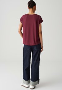 Burgunderfarbenes Kurzarmshirt kombiniert mit dunkelblauen weit geschnittenen Jeans mit umgeschlagenen Säumen und weißen Sneakers. Schlichtes, entspanntes Design.