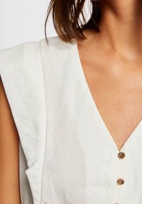Top de lino blanco con un profundo escote en V, hombros plisados y botones dorados en la parte delantera. Textura suave con mínimas ornamentaciones.
