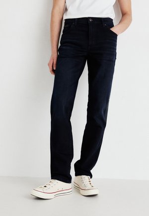 Jeans en denim bleu foncé avec une coupe droite, un design à cinq poches et une fermeture à boutons classique, associés à des baskets de couleur crème.