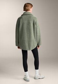 Overdime groene fleece jas met hoge kraag, gestructureerd oppervlak en ronde zoom, gecombineerd met zwarte leggings en lichte sneakers.