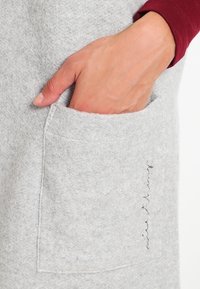 Šedá plátěná kapsa s viditelným šitím a rukou, která do ní sahá; měkká textura, ležérní design a minimalistické detaily.