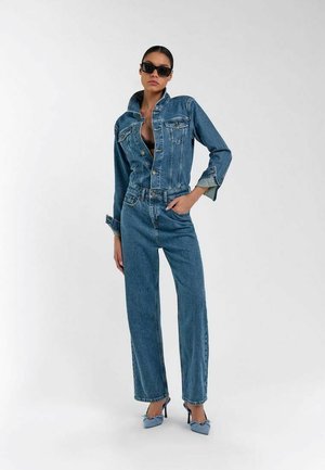 Donna in piedi che indossa giacca di jeans blu e jeans, occhiali da sole neri, tacchi alti con fiocchi, mano sinistra in tasca, su sfondo bianco.